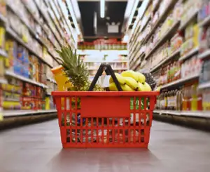 A Red Shopping Basket Filled With Groceries, Including A Pineapple, Bananas, And A Bottle Of Juice, Sits In The Middle Of A Supermarket Aisle.