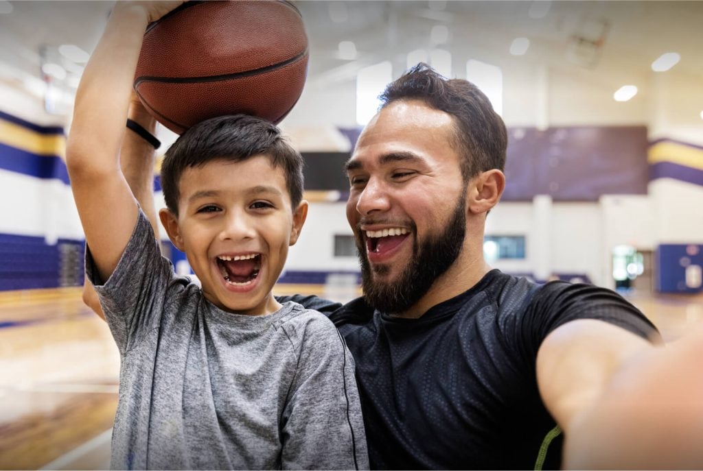 A father and son on basketball court, smiling at the camera.