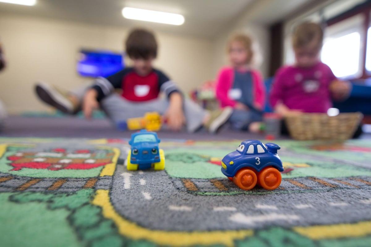 Toy cars on a play carpet are in the foreground. Three young children are blurry in the background.