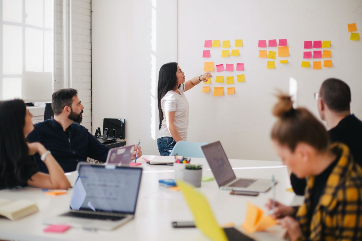 People sit around a table with laptops. One person is at a whiteboard pointing to sticky notes.