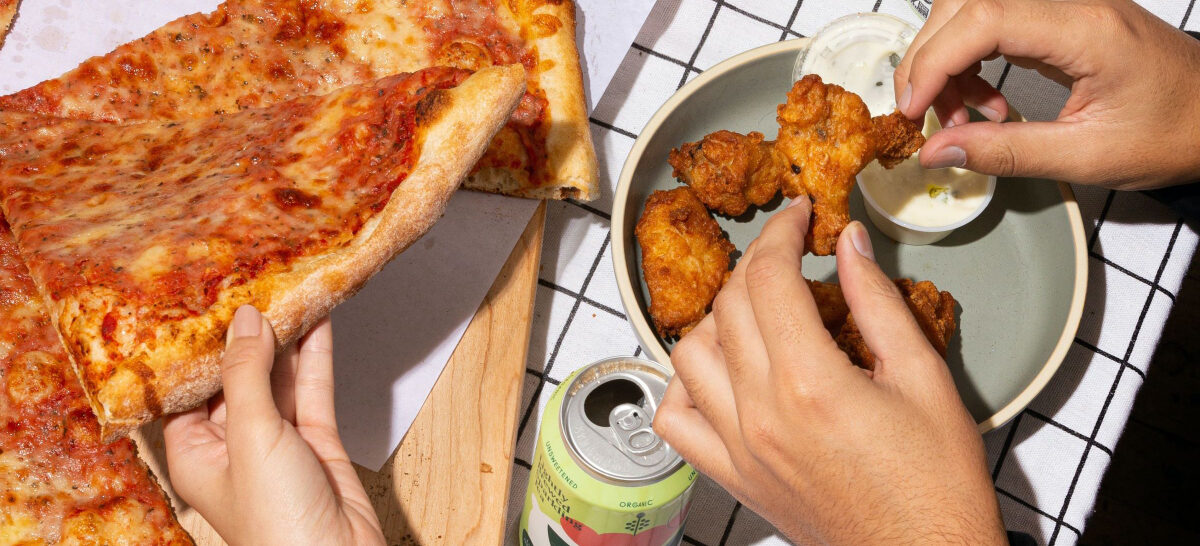 A table with a spread of food and a hand grabbing a slice of cheese pizza while two hands grab fried chicken wings.