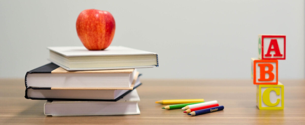 A school desk with a stack of books, an apple, colored pencils and 'ABC' blocks.