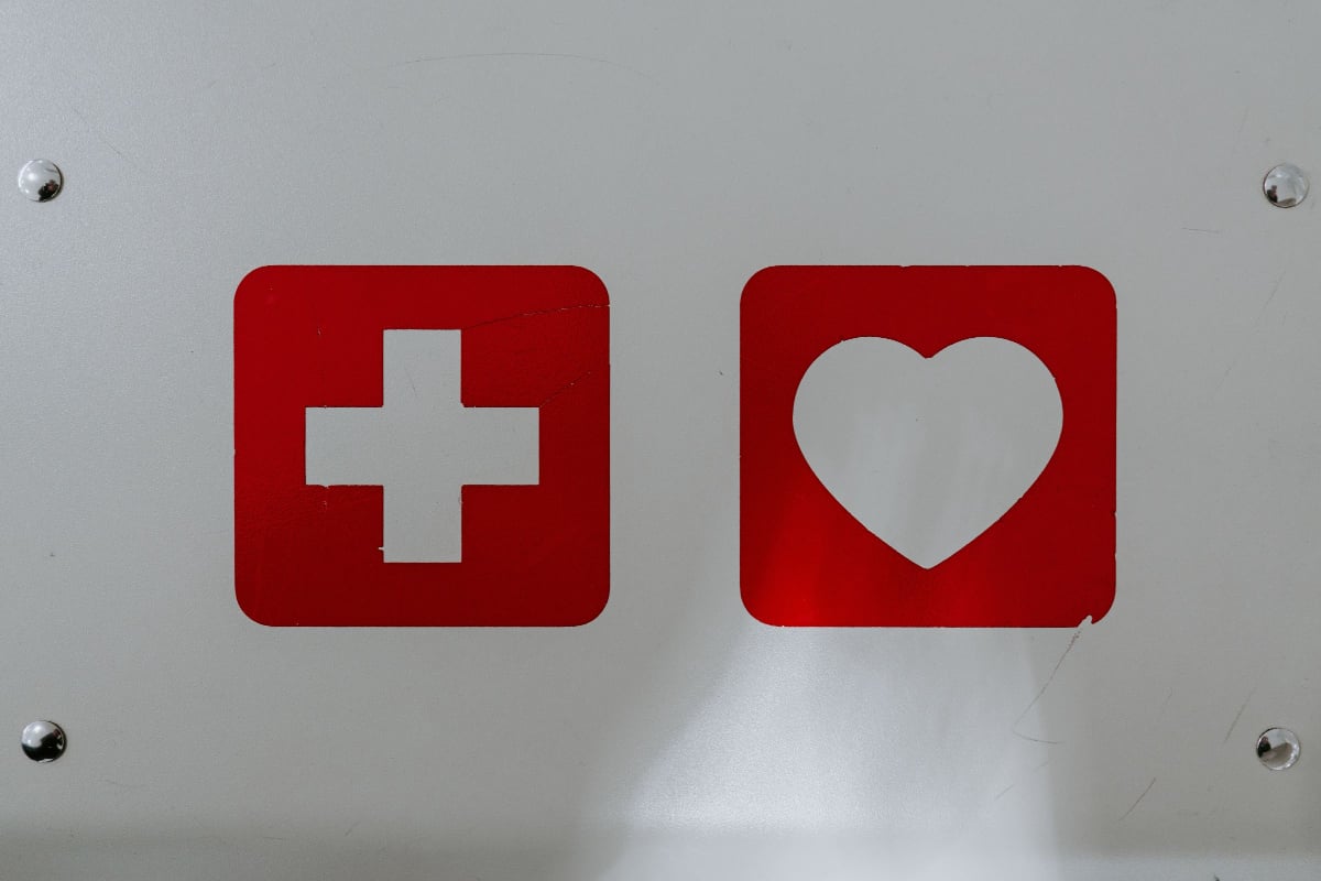 A white wall has two red signs on it, one with a white plus sign and one with a white heart.