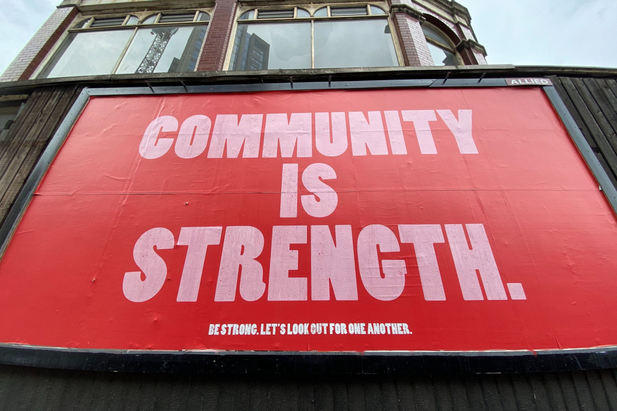 A red sign on the side of a building reads: Community is Strength. Be Strong. Look Out for One Another.