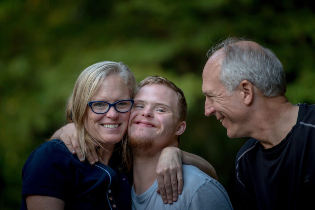 Mom hugs adult son with with down syndrome while dad smiles at them.
