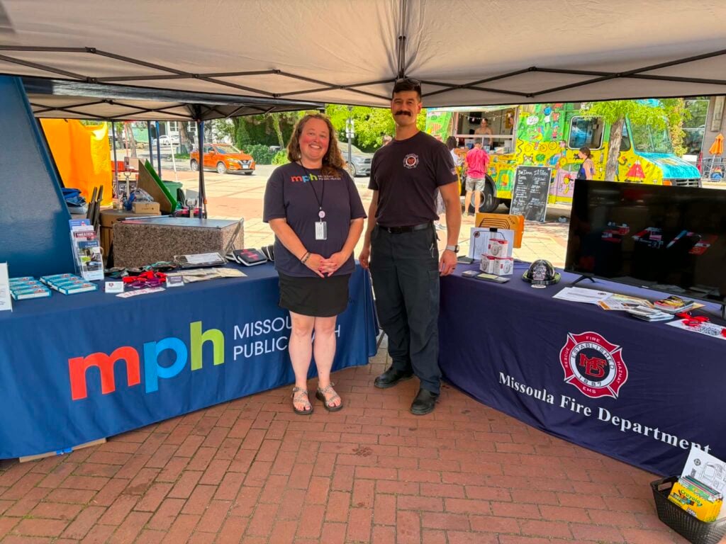 Two people stand in front of two outreach tables at Missoula's Out to Lunch event held earlier this week. One is a firefighter with the Missoula Fire Department, and the other is a staff member from Missoula Public Health. The tables are covered with educational materials about CPR, overdose recognition and response, and naloxone